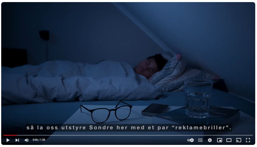 Skjermbilde fra videoen "Se verden med reklamebriller". Bildet viser en gutt som ligger og sover. På nattbordet i forgrunnen ligger et par briller. Teksten nederst på skjermen lyder: "så la oss utstyre Sondre her med et par "reklamebriller"."