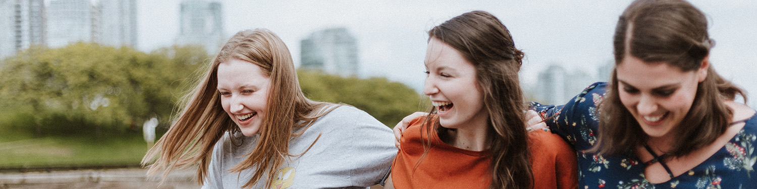 Foto av tre unge jenter som holder hverandre om skuldrene. I bakgrunnen ser vi blokker og en park.